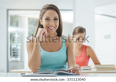 Happy smiling female student in the classroom sitting at desk and reading a book, she is looking at camera