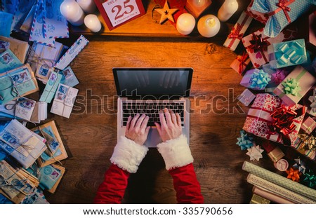Santa working at desk and typing on a laptop surrounded by colorful Christmas gifts and letters, hands top view