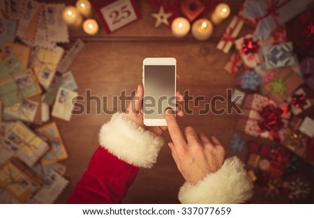 Santa Claus using a touch screen smart phone, hands close up, top view, desktop with gifts and Christmas letters on background