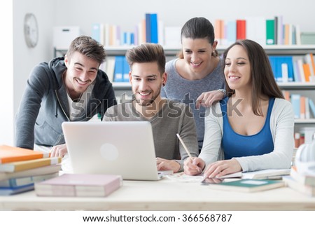 Group of smiling college students using a laptop and studying together in the classroom, education and friendship concept