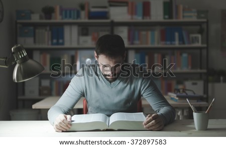 Smart confident young man studying late at night, he is sitting at desk and reading a book, knowledge and learning concept