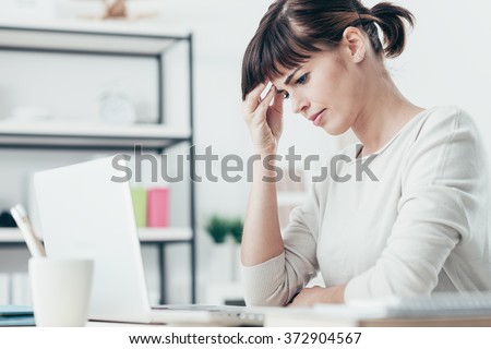 Sad tired woman having a bad headache, she is sitting at office desk and touching her temple