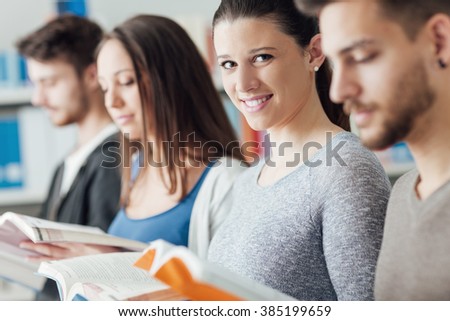 Group of college students in the library standing in line, holding books and reading, one is smiling at camera