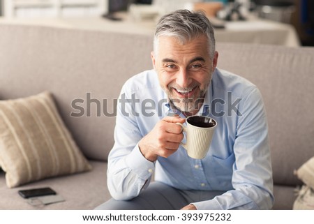 Man relaxing at home on the couch and having a coffee break, he is smiling at camera and holding a cup