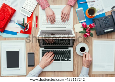 Coworkers sharing the same desk with customized workspace and working with a laptop, top view