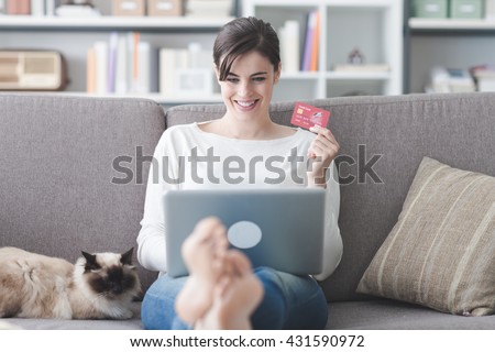 Young smiling woman at home, she is relaxing on the couch with her cat and shopping online using a credit card