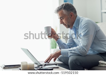Businessman relaxing at home in the living room, he is sitting on the sofa, having a coffee and working with a laptop