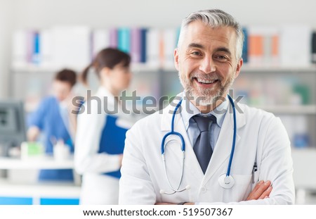 Smiling doctor posing with arms crossed in the office, he is wearing a stethoscope, medical staff on the background