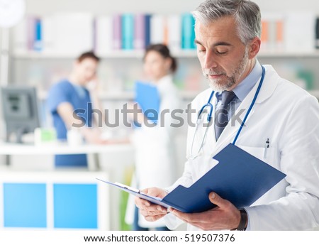 Confident professional doctor in the office checking patient\'s medical records on a clipboard, medical staff working on the background