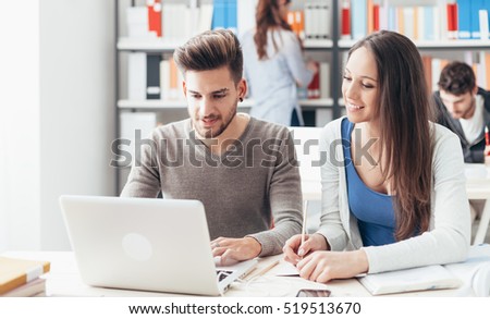 Smiling college students sitting at desk and studying together, they are using a laptop and connecting to internet, friendship and education concept