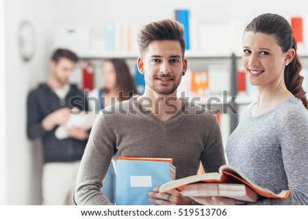 Smiling schoolmates holding books and studying together in the school library, learning and education concept