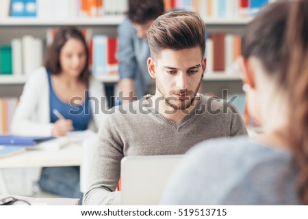 Male student sitting at desk and using a laptop, students and bookshelves on background