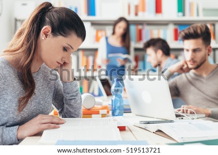 College students sitting at desk and studying together using laptops, a girl is reading a book on foreground