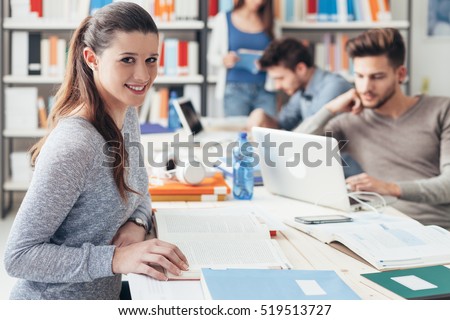 College students sitting at desk and studying together, reading books and using laptops, smiling girl on foreground