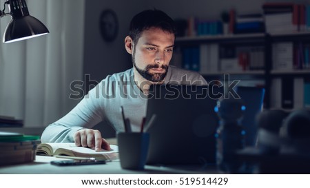 Young man sitting at desk, using a laptop, working late at night, he is studying a book and using a laptop