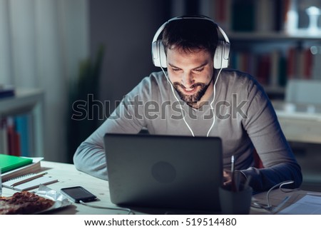 Young smiling man networking and connecting to internet using a laptop late at night, he is sitting at desk and wearing headphones