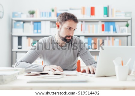 Young handsome man sitting at office desk, studying a book and connecting to internet using a laptop