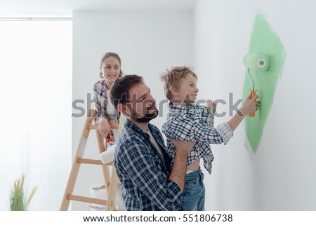 Happy young family renovating their home, the father is holding his son and he is helping him to paint a wall with a paint roller, the mother is standing on the ladder and smiling