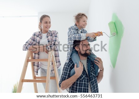 Happy family renovating their new home, the father is piggybacking his son and helping him painting with a roller, the mother is smiling and standing on a ladder
