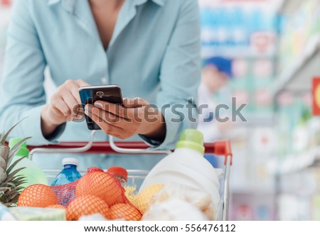 Woman doing grocery shopping at the supermarket, she is leaning on the shopping cart and connecting with her phone, apps and retail concept