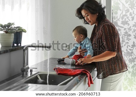 Mother is reading digital tablet while holding her baby on hands  and trying to do some home work in her kitchen.