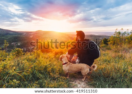 Man with beard and his small yellow dog enjoying mountain sunset and looking at the distance. He is waring black backpack and black sport sweater.