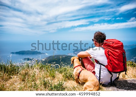 Man with red backpack and small yellow dog sitting on a mountain and looking at sea horizon