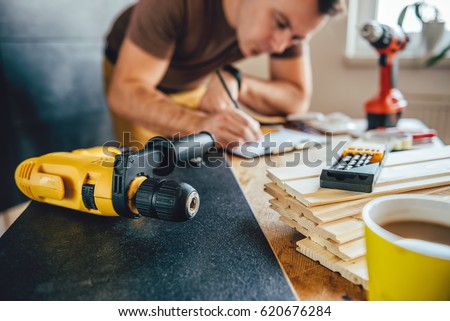 Yellow power drill and man making draft plan using pencil on the table with tools in the background