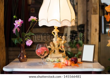Antique lamp with cherubs on a side table with flowers in a vase and a picture frame with a blank white center for your portrait