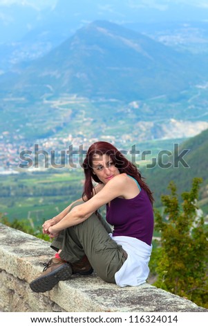 Woman wearing a tank top and pants,  indian sitting on a ledge overlooking the city, her arms around her knees  with mountains visible in the background.