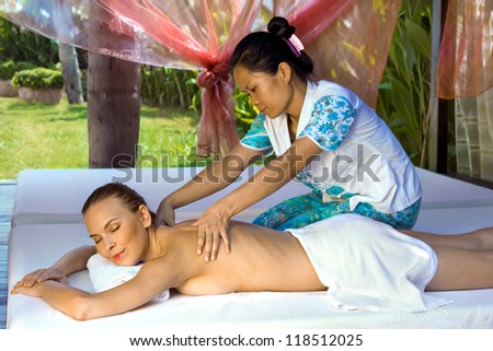 Woman in a prone position, getting a back massage from a thai masseuse inside a spa hut, grass and trees visible in the background.