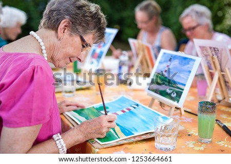 Art therapy for senior ladies, group of women painting the picture of lighthouse from the watercolor template  sitting at one table outdoors in the park.