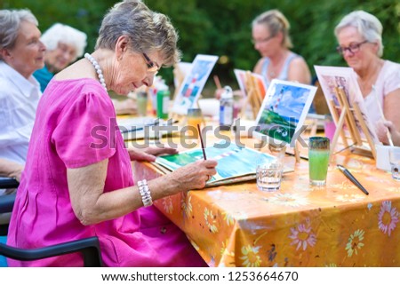 Stylish senior lady painting in art class with friends from her care home for the aged copying a painting with water colors on a canvas outdoors at a table in the garden.