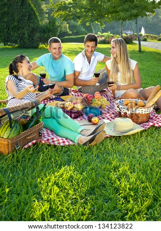 Two attractive young couples picnicking in a park relaxing on a fresh red and white cloth spread out on green grass as they chat and enjoy their wine and food