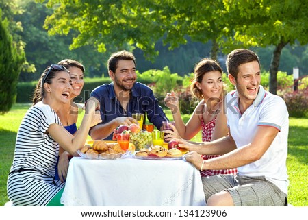 Group Of Multi Ethnic Friends Having Picnic. Group Of Adult Family Members Laughing Heartily As They Look Over Their Shoulders At Something Off Screen To The Right