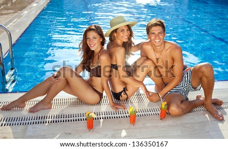 High angle view of two beautiful women in bikinis sitting sunbathing at the edge of a sparkling blue swimming pool with a handsome young man enjoying refreshing drinks