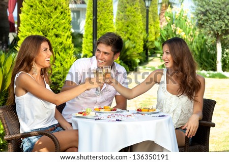 Three Friends Celebrating Seated At An Outdoor Table In The Garden Toasting Each Other With Champagne And Enjoying A Healthy Tropical Fruit Meal