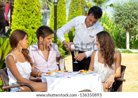 Waiter pouring wine for three young attractive friends seated at an outdoor table in the garden of a restaurant