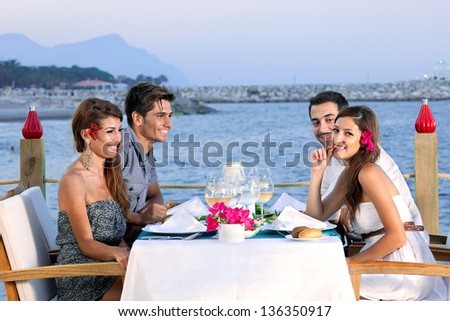 Two attractive young couples celebrating at a seaside restaurant sitting at a table at the waters edge waiting for their meal and enjoying a glass of wine