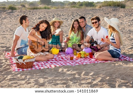 Group of happy young people having a picnic on the beach. Students having an enjoyable picnic on the beach with healthy food, some making music, young female is playing guitar.