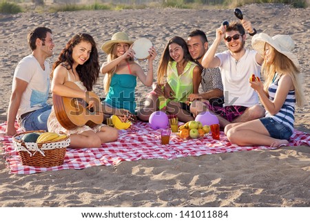 Group of happy young people having a picnic on the beach. Group of young people having an enjoyable picnic on the beach with healthy food, three of them playing music, young female is playing guitar.