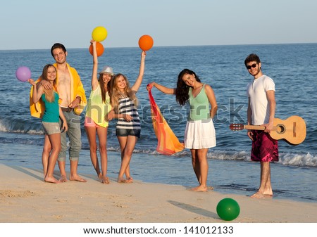 Group Of Young People Enjoying Beach Party With Guitar And Balloons. Young Friends Enjoying Beach Party With Balloons And Guitar, Standing On The Beach, Blue Sea And Sky Behind Them.