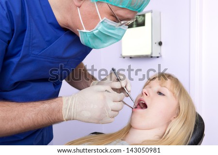 Dental-Surgeon examining the teeth of a woman. Dental-Surgeon using a mask and cap performing an examination of the teeth on a beautiful blond woman  with teeth x-ray in the back