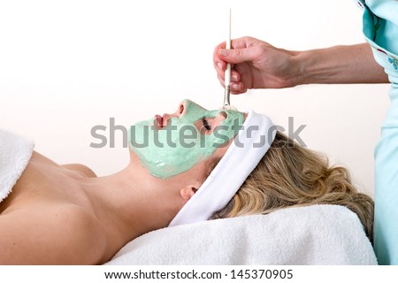 Beautician applying green thalasso facial mask with a brush on the forehead of a laying and relaxed beautiful blond woman against a white background