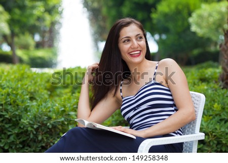 Beautiful female student is reading a book outdoors in a park, looking to somebody on her side.