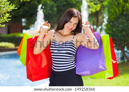Young happy female shopping, carrying lots of colorful shopping bags, in the background some fountains next to a shopping mall.