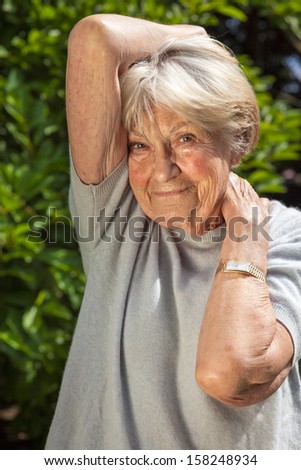 Positive confident elderly woman. Positive confident elderly woman with a warm friendly smile standing looking at the camera with her arms raised to her head and neck against green foliage