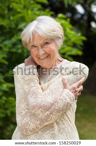 Smiling friendly senior woman. Smiling friendly senior woman in a stylish lacy blouse holding her arms around herself as she stands in a sunny summer park