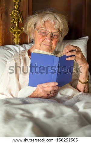 Elderly woman in her nightgown wearing glasses sitting propped up against the pillows reading a hardcover book in bed