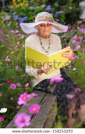 Fashionable old lady reading in her garden on a summer day sitting amongst the flowers on a wooden bench wearing sunglasses and an elegant wide brimmed sunhat